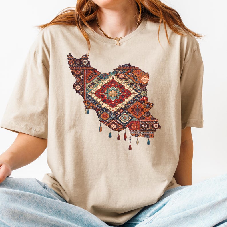 May include: Beige t-shirt featuring a detailed map of Iran filled with a colorful, patterned rug design. The rug design includes red, blue, and orange hues, with decorative tassels hanging from the bottom of the map.
