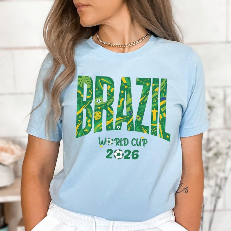 May include: Light blue t-shirt with the word "BRAZIL" in green and yellow, featuring decorative patterns. Below the word "BRAZIL" is the text "WORLD CUP 2026" with a soccer ball graphic. The shirt is a crew neck style.