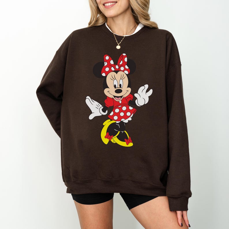 Op de afbeelding: Een bruine sweatshirt met een Minnie Mouse-ontwerp. De afbeelding toont Minnie Mouse in een rode jurk met witte stippen, een bijpassende strik en gele schoenen. De sweatshirt heeft een witte kraag.