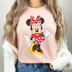 May include: A light pink t-shirt featuring a cartoon illustration of Minnie Mouse. Minnie Mouse is depicted in a red dress with white polka dots, a matching bow, and yellow shoes. The character is smiling and waving.