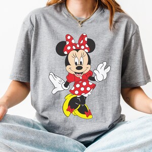 May include: A gray t-shirt featuring a Minnie Mouse graphic. Minnie is depicted in her classic outfit: a red dress with white polka dots, a matching bow, and yellow shoes. The design is centered on the shirt.