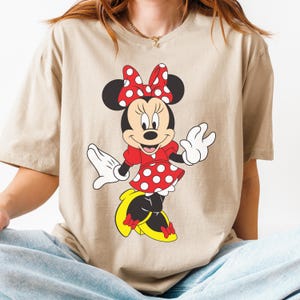 May include: Beige t-shirt featuring a cartoon illustration of Minnie Mouse. Minnie is depicted in a red dress with white polka dots, a matching bow, and yellow shoes. The shirt is a neutral color.