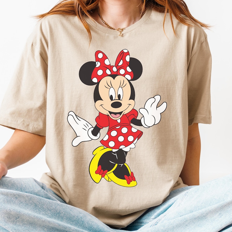 May include: Beige t-shirt featuring a cartoon illustration of Minnie Mouse. Minnie is depicted in a red dress with white polka dots, a matching bow, and yellow shoes. The shirt is a neutral color.