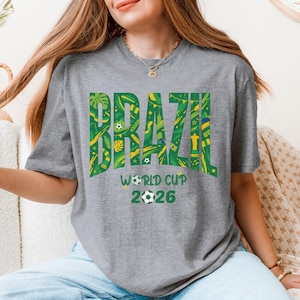 May include: A gray t-shirt with the word "BRAZIL" in large green letters filled with yellow and green patterns. Below the word "BRAZIL" is the text "WORLD CUP 2026" with a soccer ball graphic.