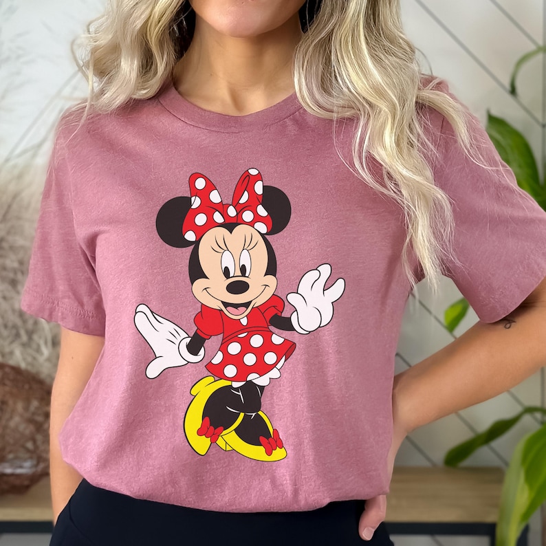 May include: A mauve-colored t-shirt featuring a graphic of Minnie Mouse. Minnie is depicted in her classic attire: a red dress with white polka dots, a matching bow, and yellow shoes. Her black ears and white gloves are also visible.