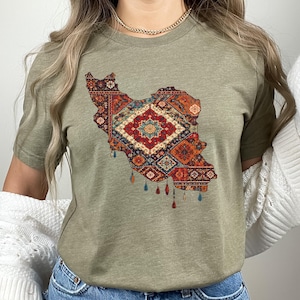 May include: Olive green t-shirt featuring a detailed map of Iran filled with a colorful, intricate rug design. The rug design includes shades of red, blue, orange, and cream, with decorative tassels hanging from the bottom of the map.
