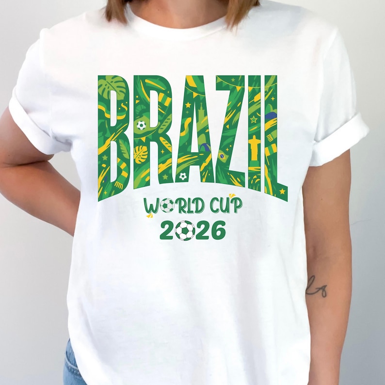 May include: White t-shirt with the word "BRAZIL" in large green letters filled with a tropical pattern. Below, the text "WORLD CUP 2026" is displayed with soccer ball graphics, celebrating the upcoming tournament.