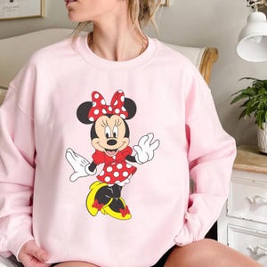 Op de afbeelding: Een lichtroze sweatshirt met een afbeelding van Minnie Mouse. Minnie draagt een rode jurk met witte stippen, een bijpassende strik en gele schoenen. Het stripfiguur lacht en zwaait.