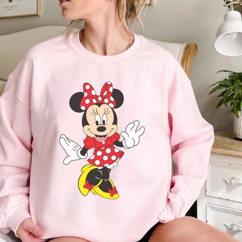 Op de afbeelding: Een lichtroze sweatshirt met een afbeelding van Minnie Mouse. Minnie draagt een rode jurk met witte stippen, een bijpassende strik en gele schoenen. Het stripfiguur lacht en zwaait.