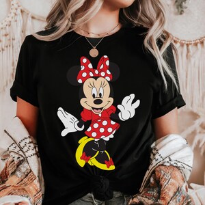 May include: Black t-shirt featuring a Minnie Mouse graphic. Minnie is depicted in a red dress with white polka dots, a matching bow, and yellow shoes. The design is a classic cartoon style.