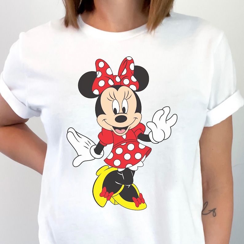 May include: White t-shirt featuring a cartoon illustration of Minnie Mouse. Minnie is depicted in a red dress with white polka dots, a matching bow, and yellow shoes. The design is centered on the shirt.