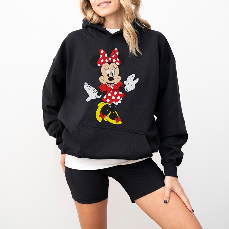 Op de afbeelding: Zwarte hoodie met een Minnie Mouse-ontwerp. De afbeelding toont Minnie Mouse in een rode jurk met stippen en een strik, met gele schoenen. De hoodie heeft een voorzak. Een wit ondershirt is zichtbaar.