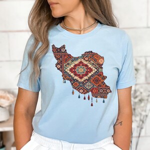 May include: Light blue t-shirt featuring a detailed map of Iran filled with a colorful, intricate rug design. The design includes shades of red, orange, and blue, with tassel-like details at the bottom. The shirt is a short-sleeved crew neck.