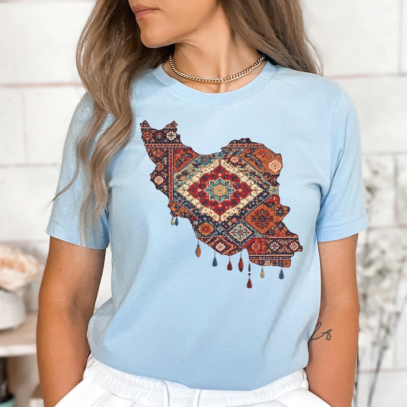 May include: Light blue t-shirt featuring a detailed map of Iran filled with a colorful, intricate rug design. The design includes shades of red, orange, and blue, with tassel-like details at the bottom. The shirt is a short-sleeved crew neck.