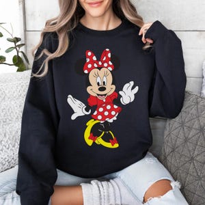 Op de afbeelding: Zwart sweatshirt met een afbeelding van Minnie Mouse. Minnie draagt een rode jurk met witte stippen, een bijpassende strik en gele schoenen. Haar witte handschoenen zijn omhoog geheven in een golf.