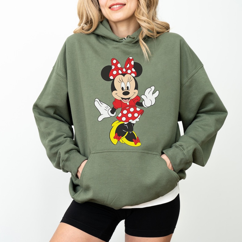 Op de afbeelding: Een olijfgroene hoodie met een Minnie Mouse-afbeelding. De afbeelding toont Minnie Mouse in een rode polkadotjurk, strik en gele schoenen. De hoodie heeft een voorzak en een wit ondershirt is zichtbaar.