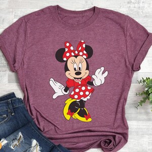 May include: Burgundy t-shirt featuring a Minnie Mouse graphic. The cartoon character is depicted in a red dress with white polka dots, a matching bow, white gloves, and yellow shoes. The shirt is paired with blue jeans.