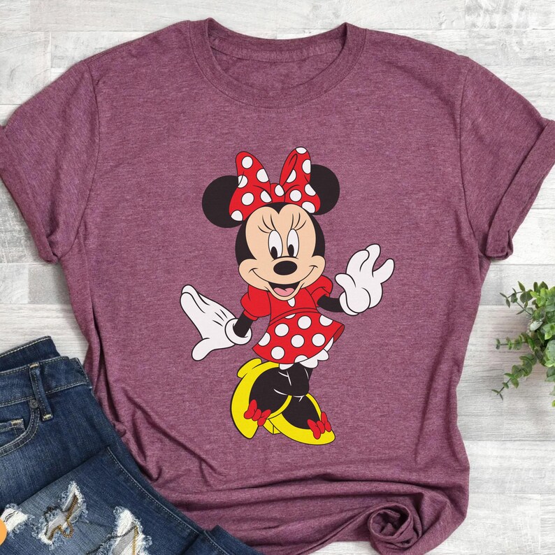 May include: Burgundy t-shirt featuring a Minnie Mouse graphic. The cartoon character is depicted in a red dress with white polka dots, a matching bow, white gloves, and yellow shoes. The shirt is paired with blue jeans.