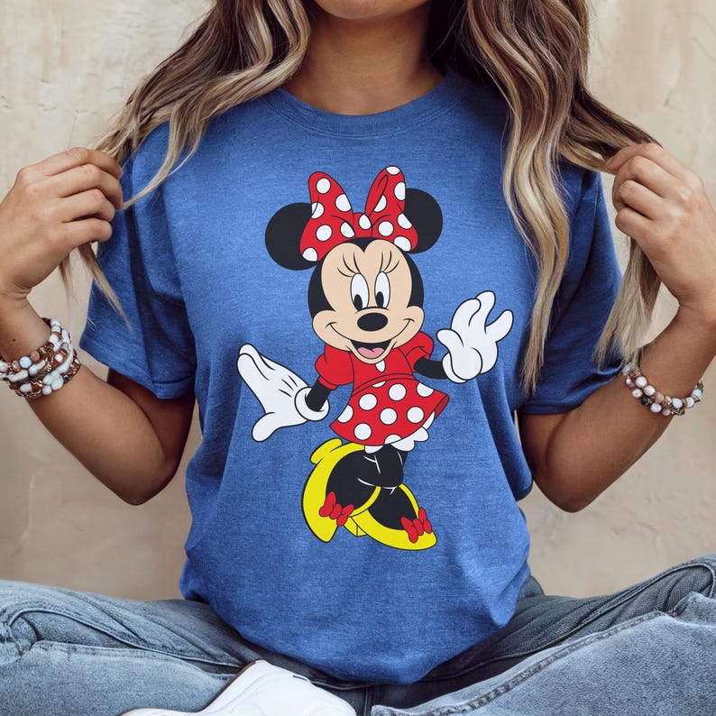 May include: Blue t-shirt featuring a Minnie Mouse graphic. The cartoon character is wearing a red dress with white polka dots, a matching bow, and yellow shoes. The design is centered on the shirt.