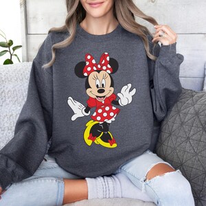 Op de afbeelding: Donkergrijze sweatshirt met een Minnie Mouse-ontwerp. Het stripfiguur is afgebeeld in een rode jurk met witte stippen, een bijpassende strik en gele schoenen. De sweatshirt heeft lange mouwen en een ronde hals.