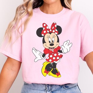 May include: A light pink t-shirt featuring a cartoon illustration of Minnie Mouse. Minnie is depicted in her classic outfit: a red dress with white polka dots, a matching bow, and yellow shoes. Her black ears and white gloves are also visible.