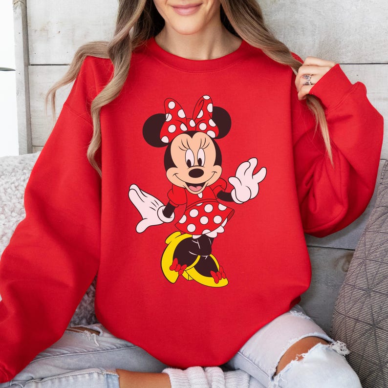 Op de afbeelding: Een rode sweatshirt met een Minnie Mouse-afbeelding. De afbeelding toont Minnie Mouse in een rode jurk met witte stippen, een bijpassende strik en gele schoenen. De sweatshirt heeft een effen rode kleur.