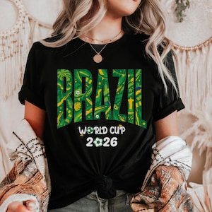 May include: Black t-shirt with the word "BRAZIL" in large green and yellow letters. The text is filled with a tropical pattern. Below, the words "WORLD CUP 2026" are printed in white, with a soccer ball graphic.