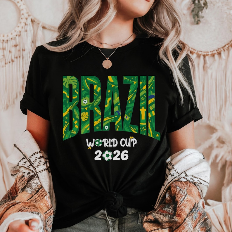 May include: Black t-shirt with the word "BRAZIL" in large green and yellow letters. The text is filled with a tropical pattern. Below, the words "WORLD CUP 2026" are printed in white, with a soccer ball graphic.