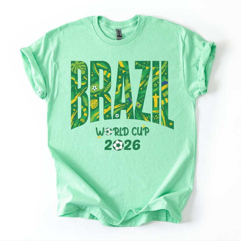 May include: A light green t-shirt with the word "BRAZIL" in large, green, patterned letters. Below, the text "WORLD CUP 2026" is displayed, with a soccer ball graphic. The shirt is a crew neck style.