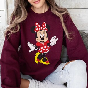 Op de afbeelding: Bordeauxkleurige sweatshirt met een Minnie Mouse-afbeelding. Minnie wordt afgebeeld in haar klassieke outfit: een rode jurk met witte stippen, een bijpassende strik en gele schoenen. Het ontwerp is gecentreerd op de voorkant van de sweatshirt.