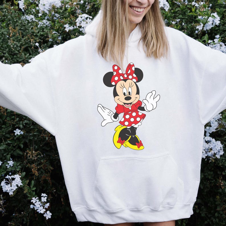 Op de afbeelding: Witte hoodie met een Minnie Mouse-afbeelding. Het stripfiguur draagt een rode jurk met witte stippen, een bijpassende strik en gele schoenen. De hoodie heeft een voorzak.