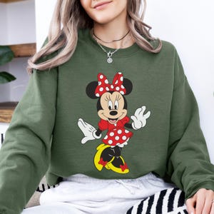 Op de afbeelding: Een olijfgroene sweatshirt met een Minnie Mouse-afbeelding. Minnie Mouse is afgebeeld in haar klassieke outfit: een rode jurk met witte stippen, een bijpassende strik en gele schoenen. De afbeelding staat in het midden van de sweatshirt.