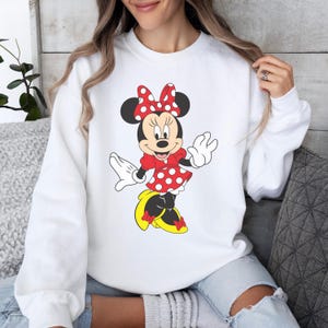 Op de afbeelding: Witte sweatshirt met een Minnie Mouse-afbeelding. Het stripfiguur draagt een rode jurk met witte stippen, een bijpassende strik en gele schoenen. Het ontwerp is gecentreerd op de voorkant van de top met lange mouwen.