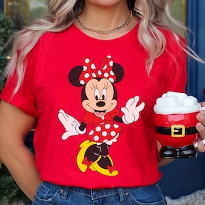 May include: A red t-shirt featuring a Minnie Mouse graphic. Minnie is depicted in her classic outfit: a red dress with white polka dots, a matching bow, and yellow shoes. A gold necklace and a Santa-themed mug are also visible.