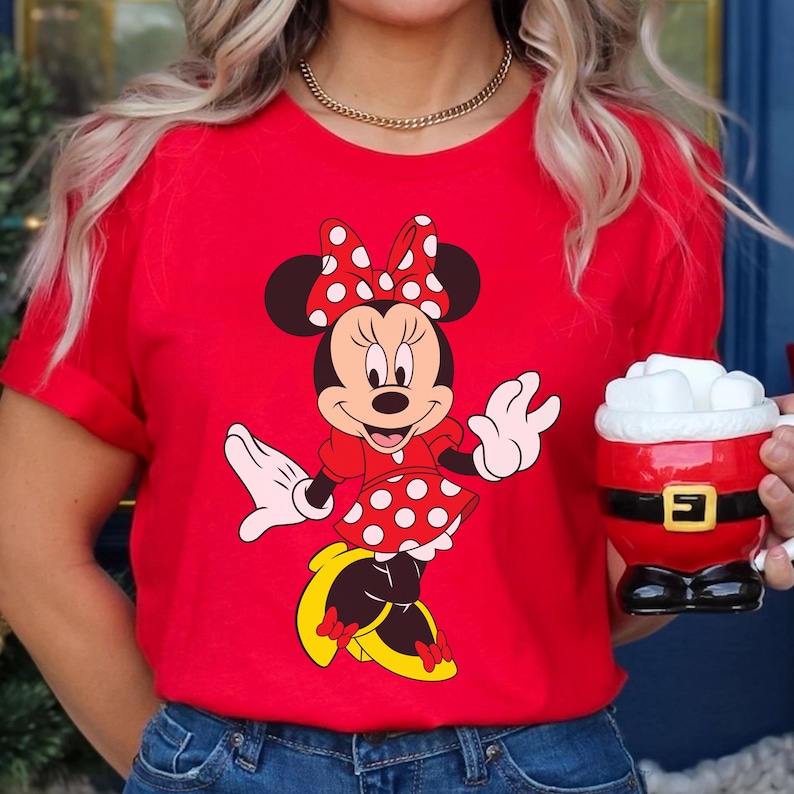 May include: A red t-shirt featuring a Minnie Mouse graphic. Minnie is depicted in her classic outfit: a red dress with white polka dots, a matching bow, and yellow shoes. A gold necklace and a Santa-themed mug are also visible.