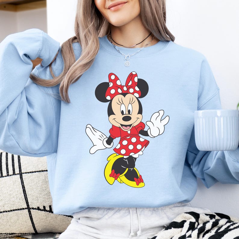 Op de afbeelding: Lichtblauw sweatshirt met een Minnie Mouse-ontwerp. Het stripfiguur draagt een rode jurk met witte stippen, een bijpassende strik en gele schoenen. De sweatshirt heeft lange mouwen.