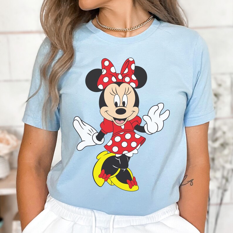 May include: Light blue t-shirt featuring a cartoon illustration of Minnie Mouse. Minnie is depicted in a red dress with white polka dots, a matching bow, and yellow shoes. The design is centered on the shirt.