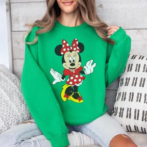 Op de afbeelding: Een groene sweatshirt met een Minnie Mouse-ontwerp. De afbeelding toont Minnie Mouse in een rode jurk met witte stippen, een bijpassende strik en gele schoenen. De sweatshirt heeft een effen, heldergroene kleur.