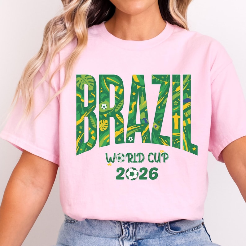 May include: A light pink t-shirt with the word "BRAZIL" in large green letters filled with yellow tropical patterns. Below, the text reads "WORLD CUP 2026" with a soccer ball graphic. The shirt is a short-sleeved crew neck.