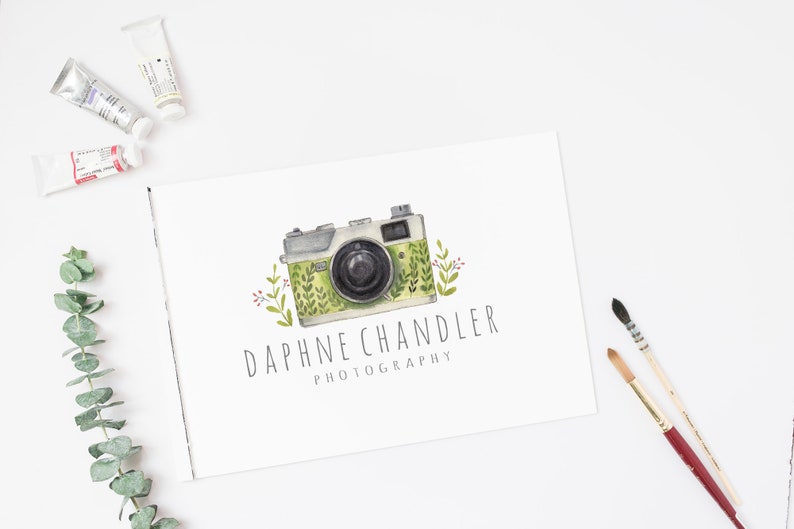 May include: A watercolor illustration of a vintage camera with green and white floral details. The text "Daphne Chandler Photography" is written below the camera.