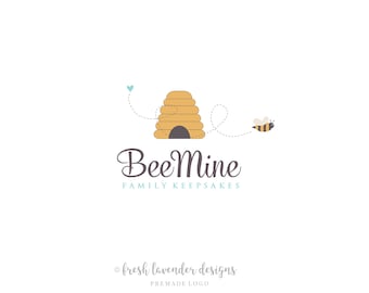 Può includere: Logo Bee Mine Family Keepsakes con un alveare giallo e marrone, un'ape che vola via, un cuore blu e il testo "Bee Mine Family Keepsakes".