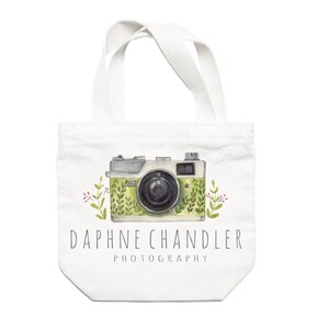 May include: White canvas tote bag with a green and white camera illustration and green foliage. The text "Daphne Chandler Photography" is printed below the camera.