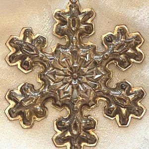 May include: A detailed, metallic snowflake ornament with six points. The snowflake has a silver-colored center and gold-colored edges. It features intricate embossed patterns and a loop at the top for hanging. This festive decoration is perfect for Christmas.