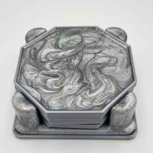 Op de afbeelding: Een set grijze onderzetters met een wervelend zilveren ontwerp, in een bijpassende houder. De onderzetters hebben een achthoekige vorm en een glanzende afwerking. De houder heeft een rechthoekige basis met gebogen zijkanten.