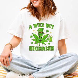 Puede incluir: Camiseta blanca con un gráfico verde de una hoja de cannabis con sombrero de copa y cerveza. El texto "A Wee Bit Highrish" está arriba y abajo del gráfico. La camiseta la lleva una persona.