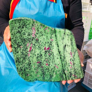 May include: A large, polished slab of zoisite mineral featuring a vibrant green base color with scattered black hornblende inclusions and distinct, elongated magenta-colored ruby crystals embedded throughout the smooth, flat stone surface.