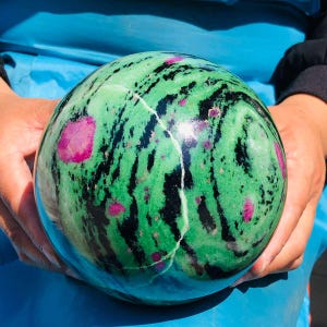 May include: A polished spherical stone featuring a vibrant green base with swirling black patterns and distinct, embedded magenta-pink crystal inclusions throughout the surface, showcasing a smooth, reflective texture and natural mineral variations.