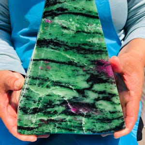 May include: A triangular slab of polished zoisite stone featuring a vibrant green base color with prominent black mineral inclusions and scattered patches of deep magenta ruby crystals throughout the surface, displaying a natural banded texture.