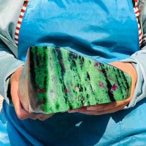 May include: A large, polished stone with a vibrant green base color, accented with black veining and small, irregular patches of deep red. The stone is held in front of a blue background, showcasing its natural beauty.