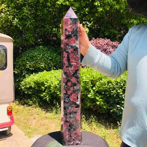 May include: A tall, six-sided crystal tower featuring a polished surface with a mottled pattern of deep red, black, and white mineral inclusions. The stone is shaped into a pointed obelisk form, displaying a smooth, reflective texture throughout.